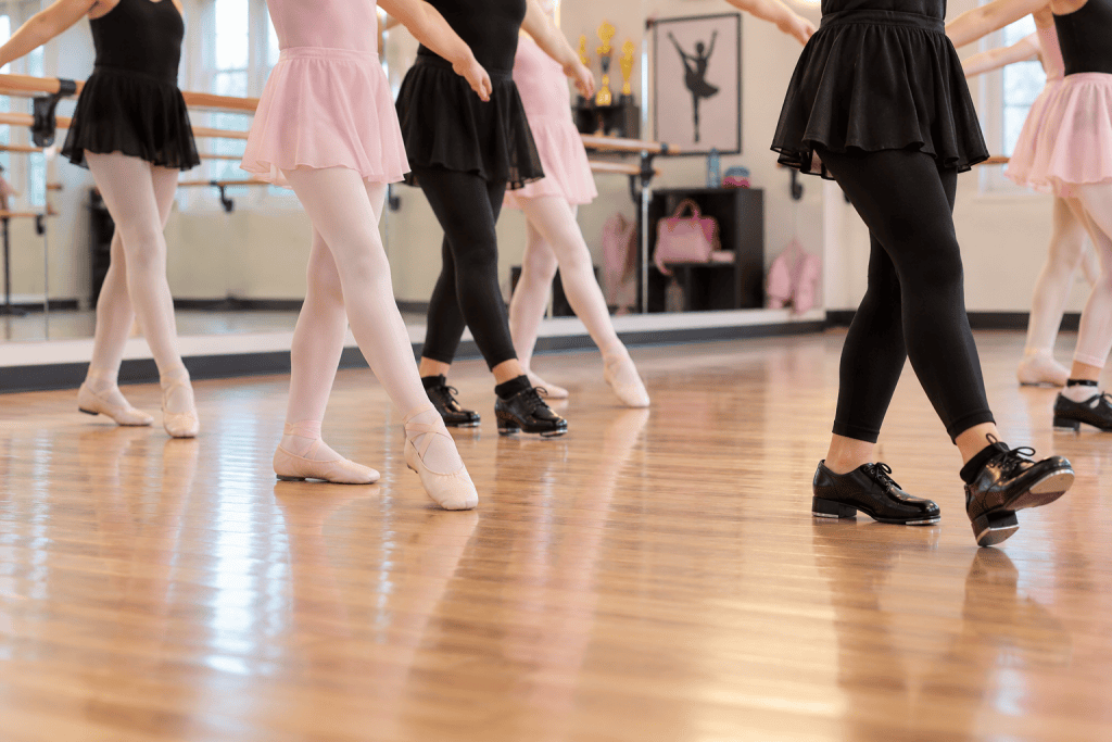 Children practicing ballet and tap dance in a studio, shown from the shoulders down, wearing ballet slippers and tap shoes on a polished wood floor.